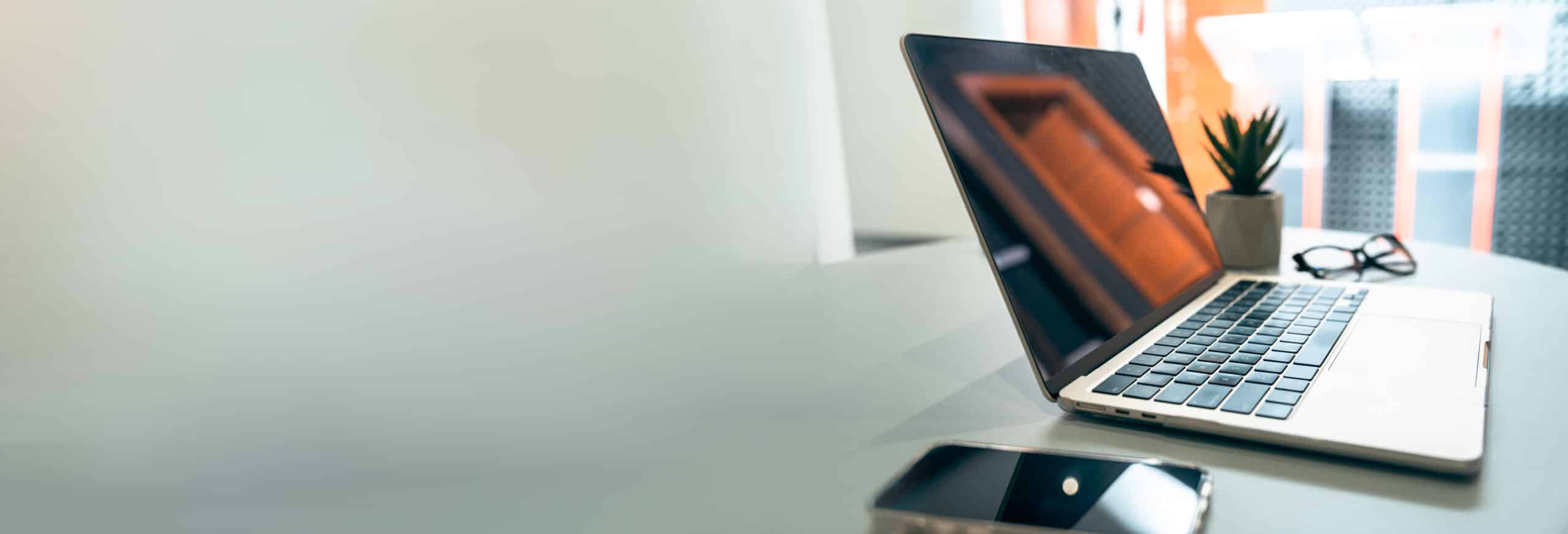 Open silver laptop on a white desk with a small plant, glasses, and a smartphone in a bright office.
