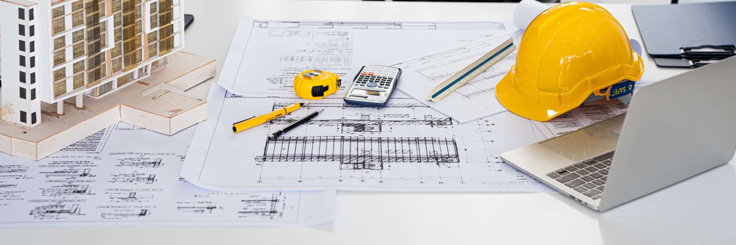 Architect or engineer professional. blueprint , structual floor plans with a pencil, triangular scale , Shop drawing , calculator, laptop and engineering tools. Architectural workspace with blueprints, a scale model building, yellow hard hat, tools, and a laptop.